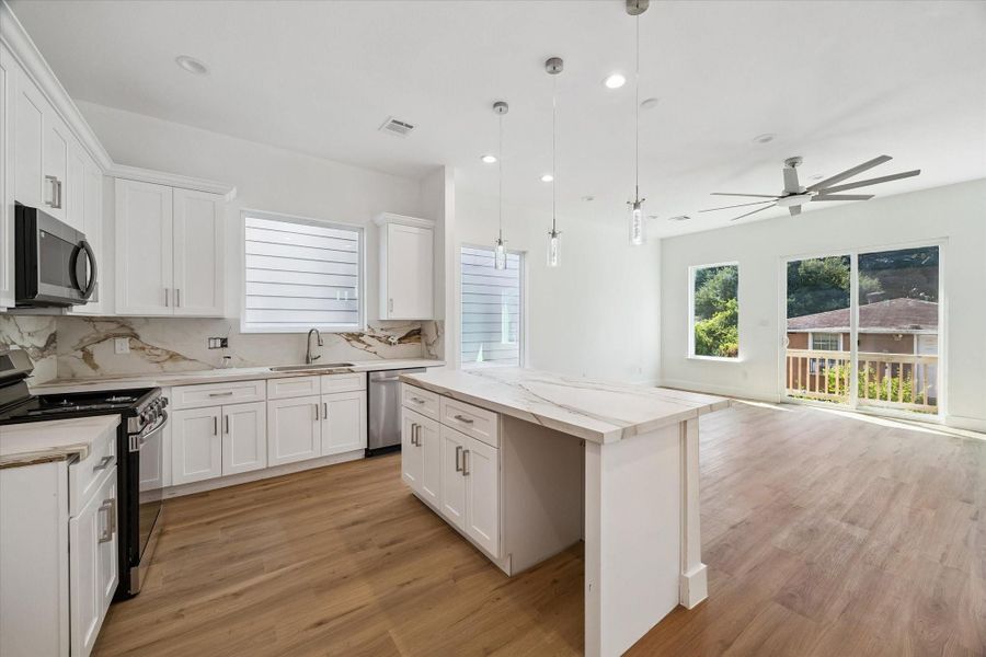 The bright kitchen opens effortlessly into the main living area, creating a contemporary space designed for both everyday living and entertaining. White cabinetry, stainless steel appliances, marble-look countertops and backsplash, pendant lighting, and warm wood-look flooring create a clean, cohesive aesthetic. A generous island provides additional prep space while large windows and sliding glass doors bring in abundant natural light.