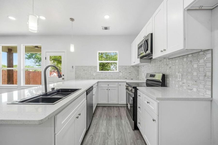 Modern kitchen featuring white cabinetry, light-toned countertops, a black double-basin sink with a gooseneck faucet, stainless steel appliances, and wood-finish flooring Modern kitchen featuring white cabinetry, light-toned countertops, a black double-basin sink with a gooseneck faucet, stainless steel appliances, and wood-finish flooring