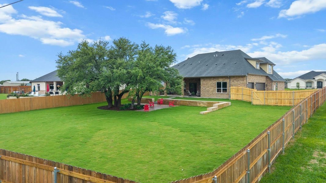 Expansive backyard featuring a manicured lawn, mature shade trees, a wooden privacy fence, a stone retaining wall, and a covered back patio