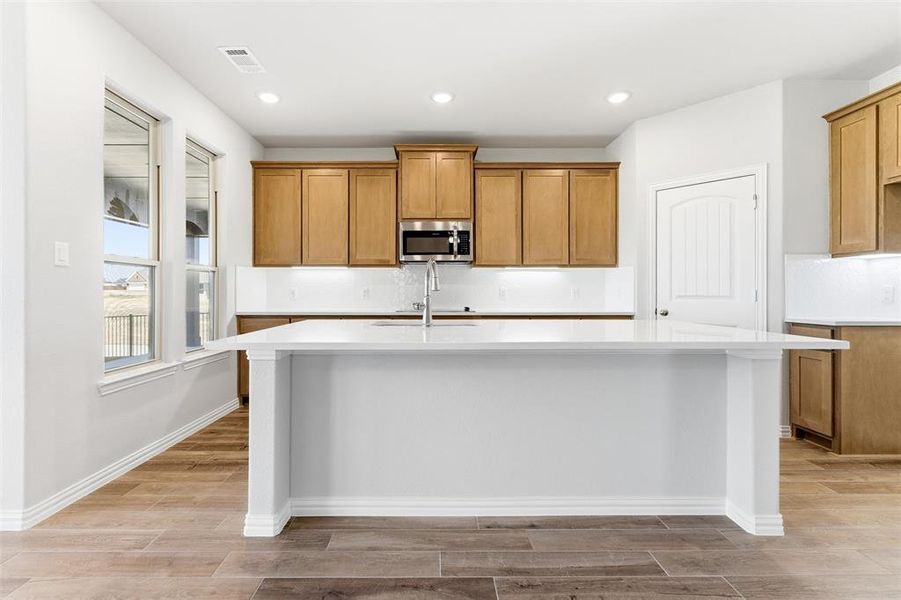 Kitchen featuring brown cabinets, an island with sink, recessed lighting, stainless steel microwave, and wood tiled floors