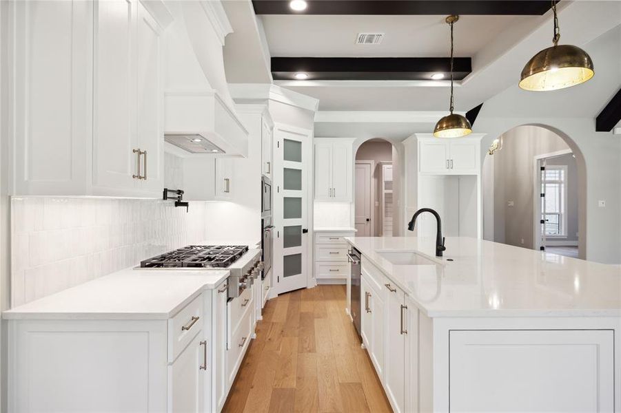 Kitchen with light wood-type flooring, custom range hood, pendant lighting, white cabinets, and an island with sink