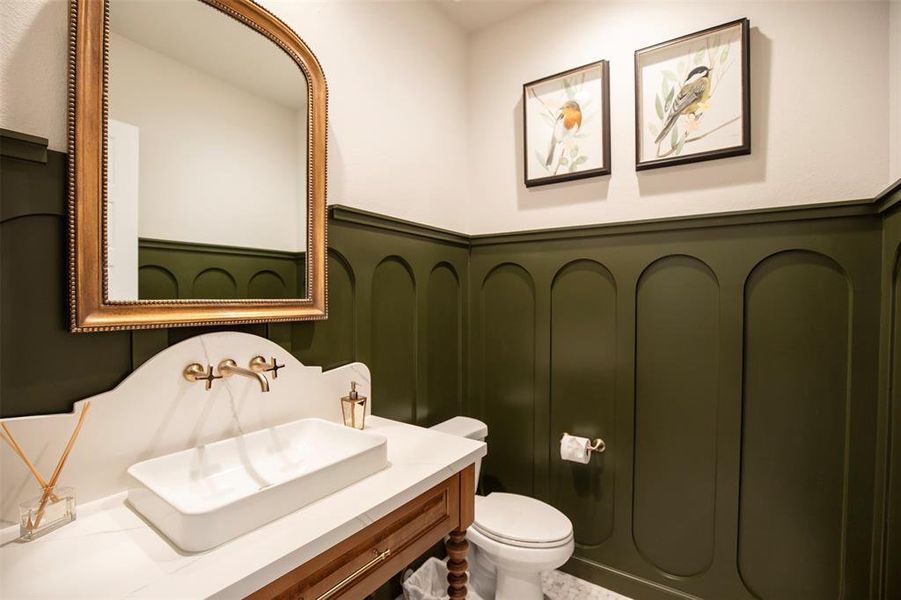 This elegant powder room features dark green wainscoting, a modern white sink with gold fixtures, and a classic wooden vanity. The space is enhanced by a large mirror and bird-themed artwork, creating a cozy and stylish atmosphere.