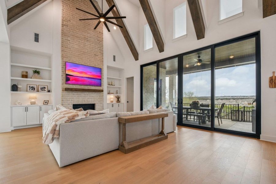 This living area features a vaulted ceiling with exposed beams and a ceiling fan, a brick fireplace, built-in shelving, and light wood flooring This living area features a vaulted ceiling with exposed beams and a ceiling fan, a brick fireplace, built-in shelving, and light wood flooring