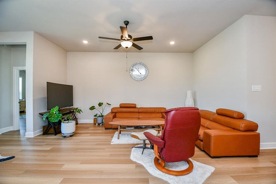 Modern living room with light wood flooring, a ceiling fan, and recessed lighting. Features a brown leather sofa set, a red recliner, and a large TV. Decorated with plants and a stylish wall clock.