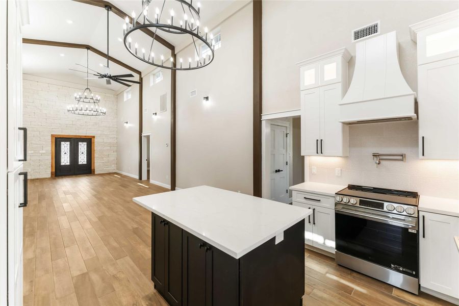 This spacious and elegant open-concept living area features high ceilings, modern chandeliers, and a stone accent wall. The kitchen boasts sleek cabinetry, a central island, and stainless steel appliances, all complemented by warm wood look tile flooring.