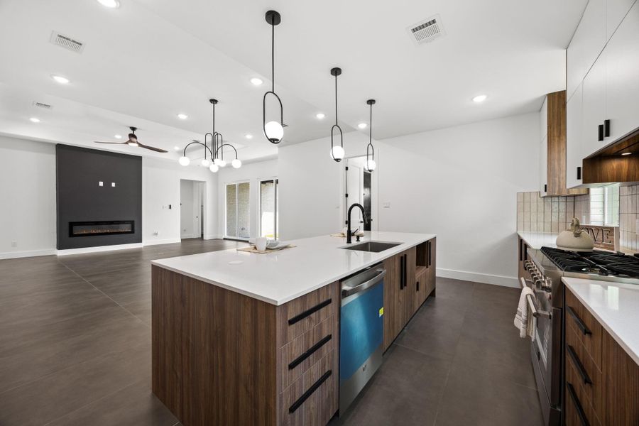 This modern kitchen and living area features an open layout with sleek finishes. The kitchen boasts a large island with pendant lighting, stainless steel appliances, and ample cabinetry. The living space includes a stylish fireplace and large windows for natural light.