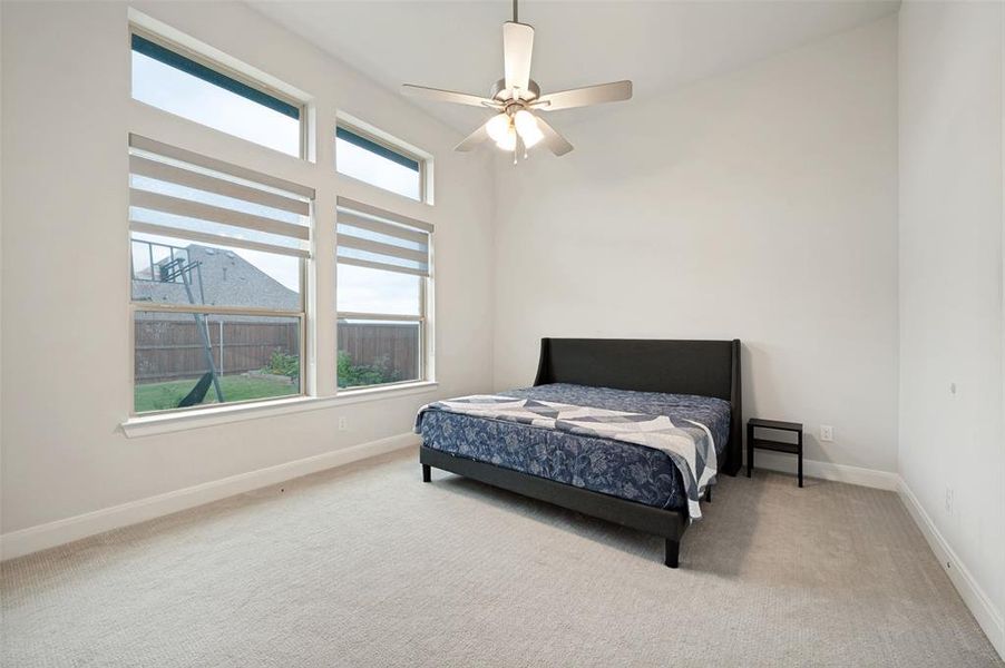 Carpeted bedroom featuring a large window with roller shades, a ceiling fan with integrated lighting, and high ceilings