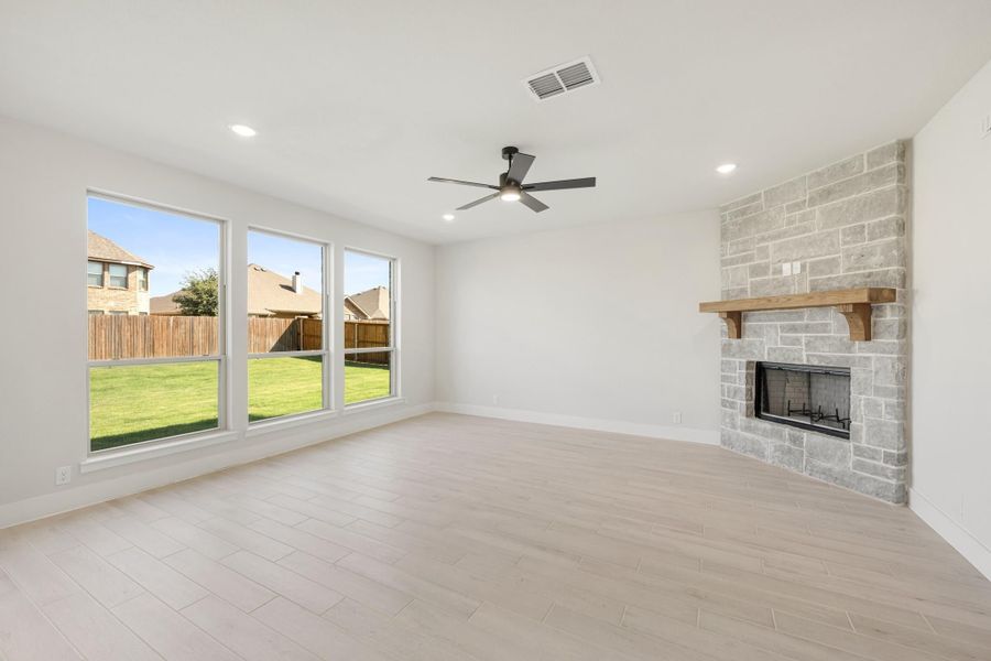 Open living room with light wood floors, stone fireplace, ceiling fan, and large backyard-facing windows