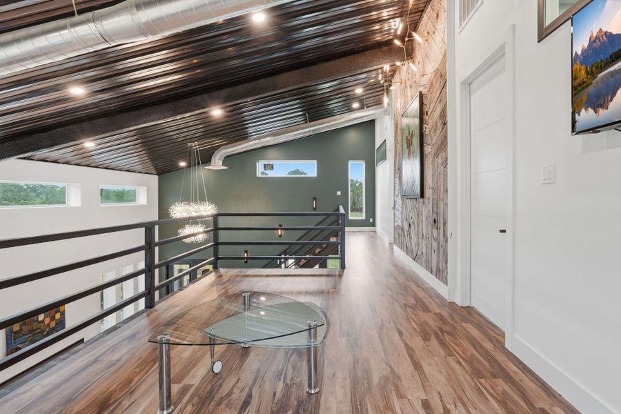 This modern loft area features a sleek metal ceiling, exposed ductwork, and stylish wood flooring. Large windows offer natural light and views, while a glass coffee table and contemporary lighting enhance the space's elegance. Perfect for a creative workspace or relaxing retreat.