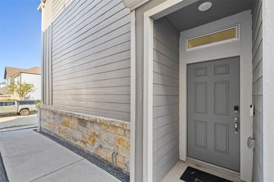 This photo showcases a modern front entrance with a gray door featuring a transom window above. The exterior has horizontal siding and stone accents along the walkway, providing a clean and inviting approach to the home.