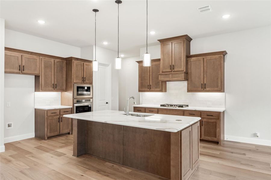 Kitchen with brown wood cabinets, large island with marble countertop, pendant lights, and light hardwood floors