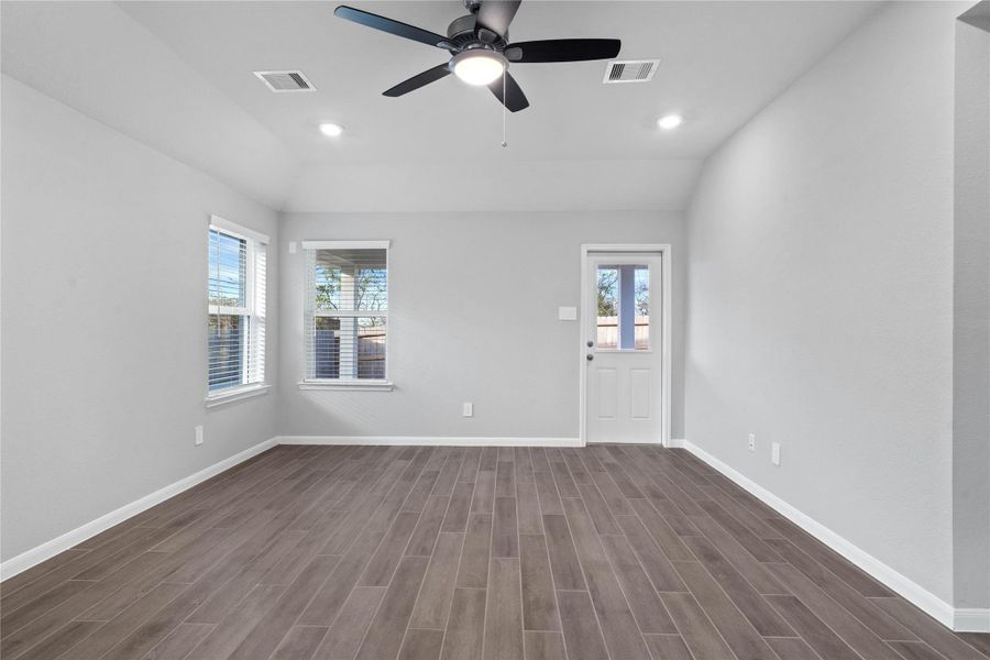A bright and airy open-concept space featuring tile flooring, sloped ceilings with recessed lighting and ceiling fan, abundant natural light from large windows (fitted with blinds), and an access door to the back covered patio.