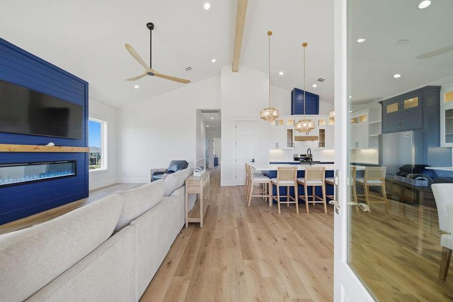 This bright, open-concept living area features a modern kitchen with blue accents, a spacious island with seating, and elegant pendant lighting. The living room boasts a cozy sectional sofa, a stylish electric fireplace, and a large wall-mounted TV. Natural wood flooring and high ceilings enhance the contemporary feel.