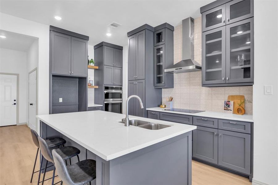 Kitchen with gray cabinetry, wall chimney exhaust hood, decorative backsplash, light wood-type flooring, and light stone counters Kitchen with gray cabinetry, wall chimney exhaust hood, decorative backsplash, light wood-type flooring, and light stone counters