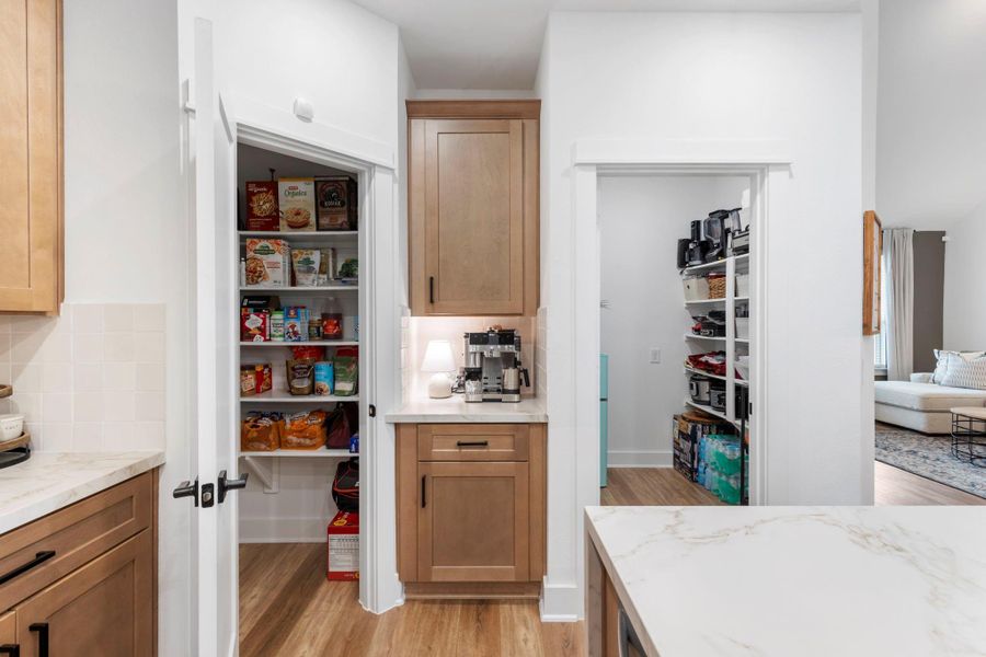 This photo showcases a bright, modern kitchen with light wooden cabinetry and a marble countertop. It features two spacious pantries, offering ample storage space for groceries and kitchen appliances. The open layout connects to a cozy living area, enhancing the home's functionality and flow.