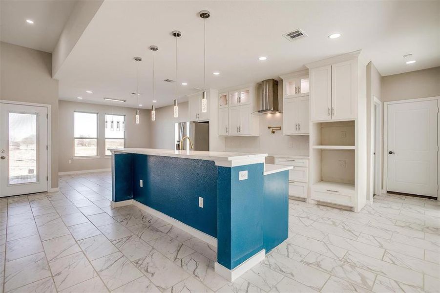 Kitchen with a center island with sink, white cabinets, wall chimney range hood, stainless steel refrigerator with ice dispenser, and decorative light fixtures