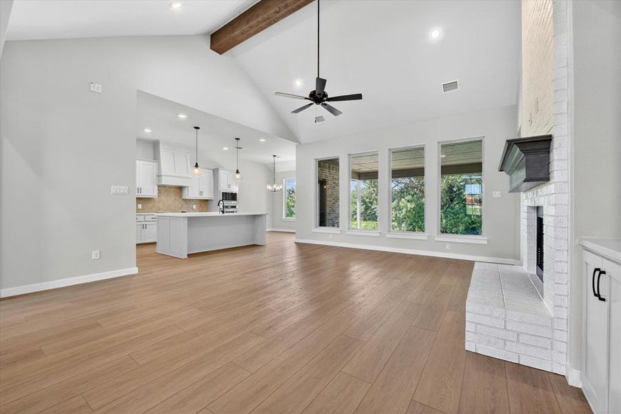Spacious open-concept living area featuring wood-finish flooring, a vaulted ceiling with exposed wood beam, and a white brick fireplace with a dark mantel