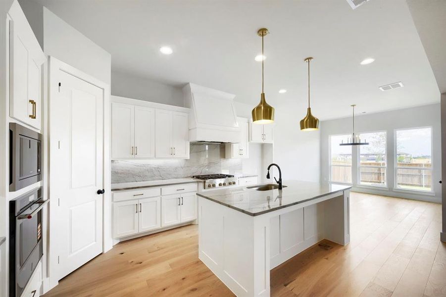 Modern kitchen featuring a central island with a sink, white cabinetry, integrated stainless steel appliances, wood-finish flooring, and ample natural light from three large windows