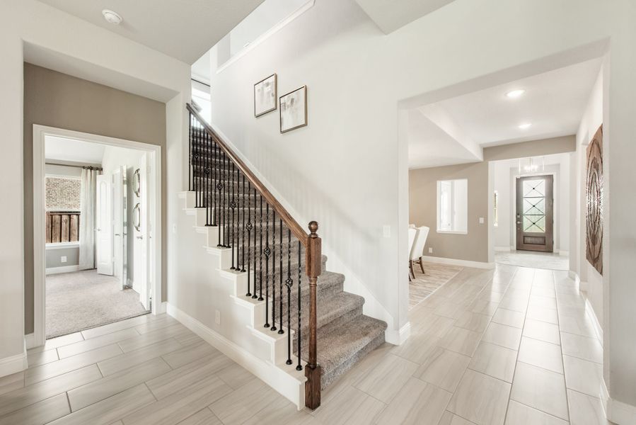 Bright home entryway with carpeted staircase, wood and iron railing, and light tile floors leading to front door.