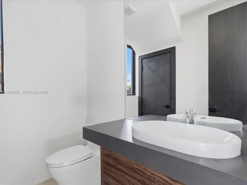 Modern floating vanity featuring a wood-finish base, dark countertop, and oval vessel sink, complemented by a large wall-mounted mirror, matte black hardware, and an elongated toilet Modern floating vanity featuring a wood-finish base, dark countertop, and oval vessel sink, complemented by a large wall-mounted mirror, matte black hardware, and an elongated toilet