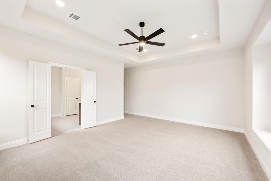 Empty bedroom with tray ceiling, black ceiling fan, carpet flooring, and open doors to ensuite bathroom