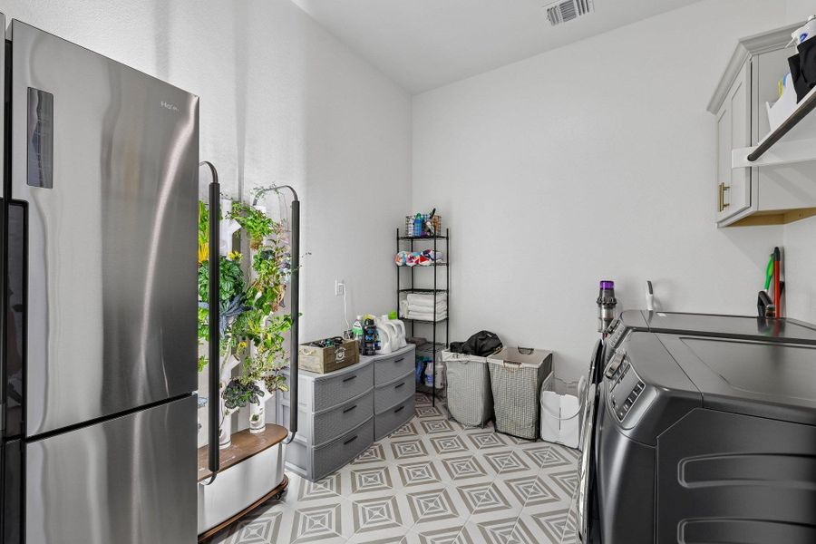 Utility room featuring a patterned tile floor, overhead cabinetry with gold-toned hardware, and a stainless steel appliance