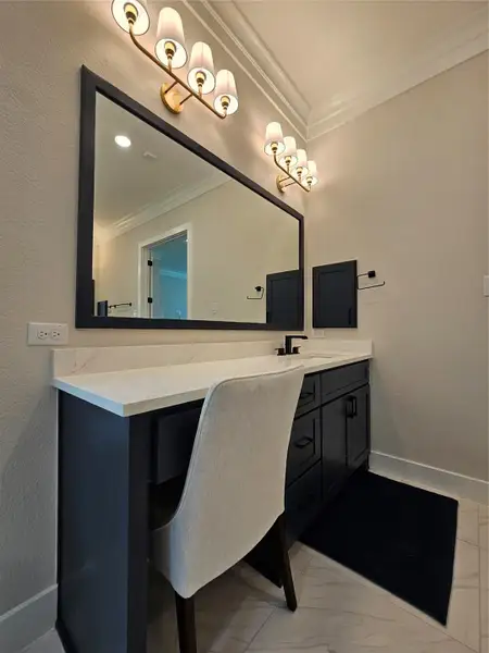 This photo showcases a modern bathroom vanity area with a large mirror and elegant lighting fixtures. The white countertop contrasts with dark cabinetry, and a cushioned chair adds comfort. The space combines functionality with style, ideal for a contemporary home.
