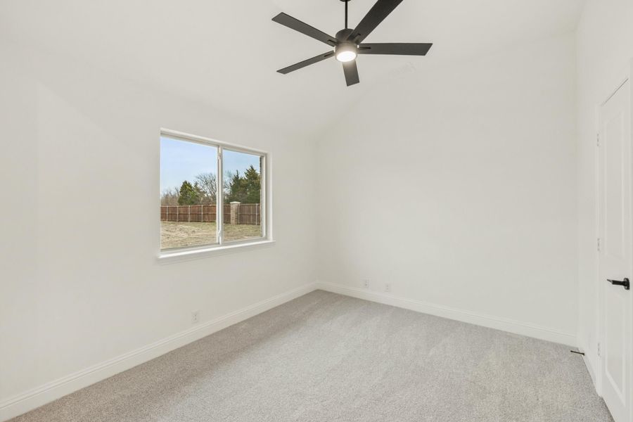 Empty bedroom with vaulted ceiling, black ceiling fan, carpet flooring, and a window overlooking a fenced backyard