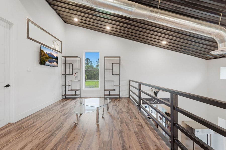 This photo showcases a modern loft area with sleek wooden flooring, industrial-style exposed ductwork, and a sloped ceiling. It features a glass coffee table, wall-mounted TV, and open shelving, with a large window offering natural light and views of greenery.