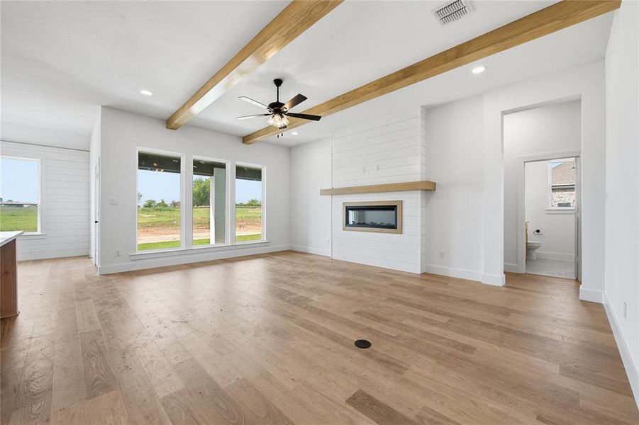 Unfurnished living room with beamed ceiling, ceiling fan, a fireplace, light wood-type flooring, and healthy amount of natural light Unfurnished living room with beamed ceiling, ceiling fan, a fireplace, light wood-type flooring, and healthy amount of natural light
