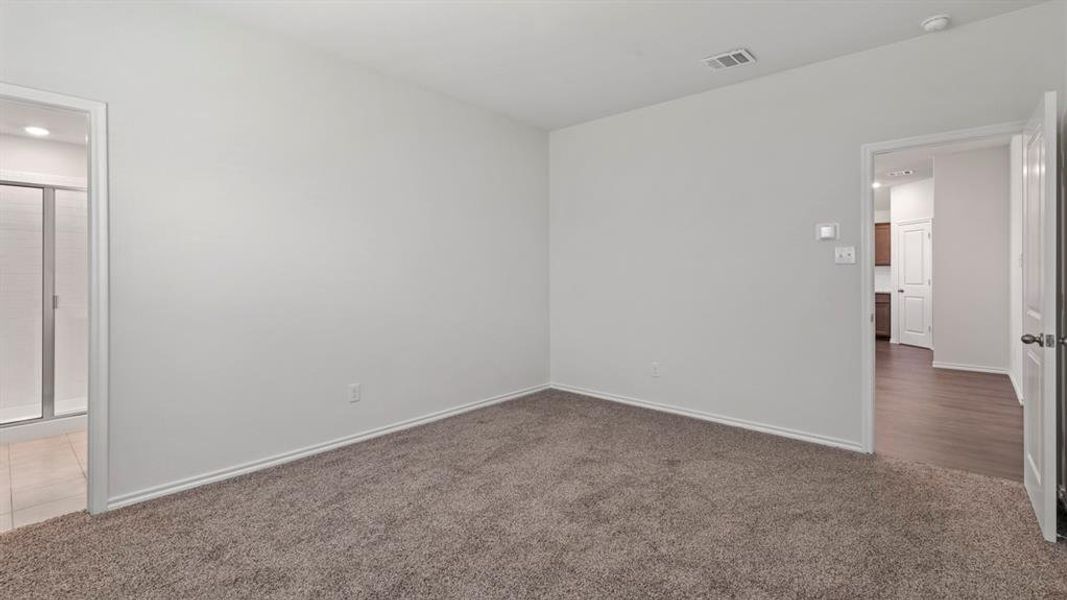 Neutral-toned carpeting throughout a spacious room with white walls, featuring an entryway to a bathroom with tiled flooring and a glass-enclosed shower, and a doorway opening to wood-finish flooring