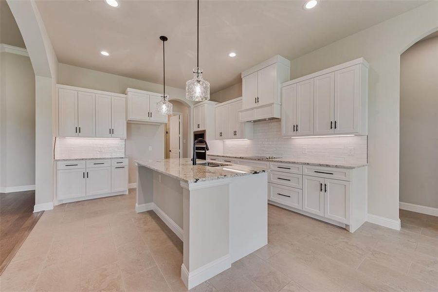 Kitchen featuring arched walkways, backsplash, light stone countertops, white cabinetry, and a center island with sink