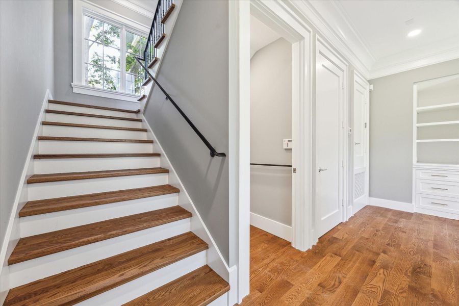 This photo showcases the modern, bright hallway with elegant hardwood flooring and a staircase featuring wooden treads and white risers. The door is to the elevator ready closet.  The space is well-lit with natural light.