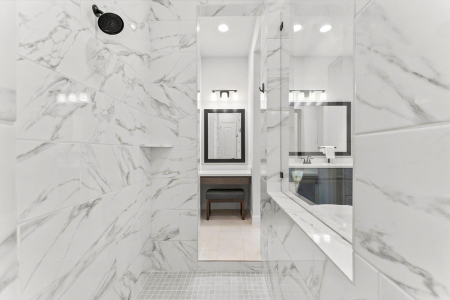 This image is taken from inside the shower, looking out into the rest of the primary bathroom. It highlights the white marble-style tile walls and the glass shower enclosure. The reflection in the mirror shows the opposite side of the bathroom with a vanity and a makeup table. This image is taken from inside the shower, looking out into the rest of the primary bathroom. It highlights the white marble-style tile walls and the glass shower enclosure. The reflection in the mirror shows the opposite side of the bathroom with a vanity and a makeup table.