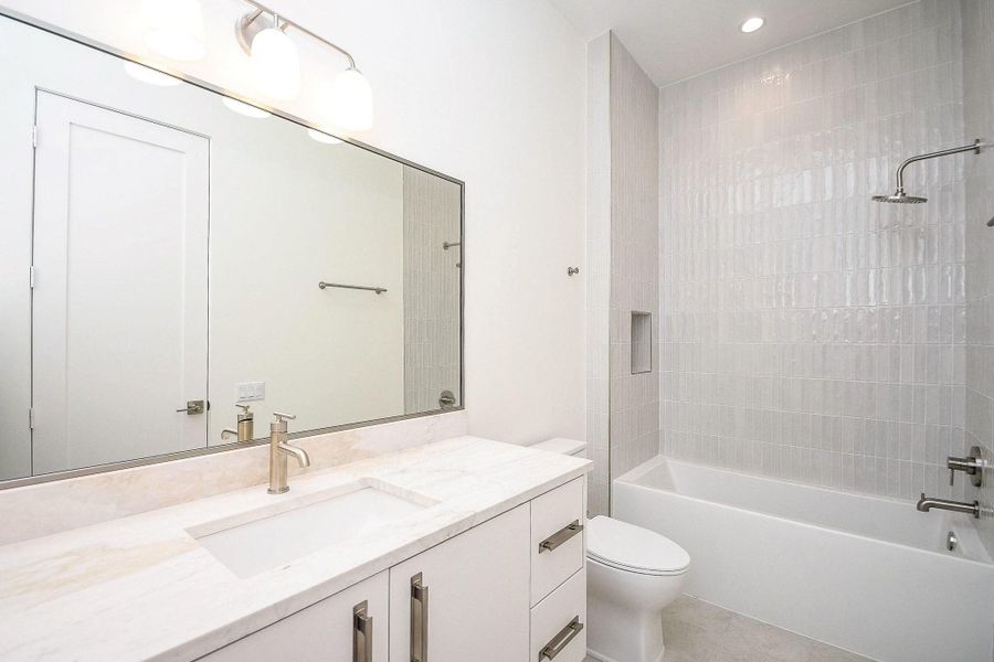 This photo showcases a modern bathroom featuring a sleek white vanity with a marble countertop, large mirror, and stylish lighting. It includes a tiled shower and bathtub combination with a chrome showerhead, creating a bright and clean atmosphere.