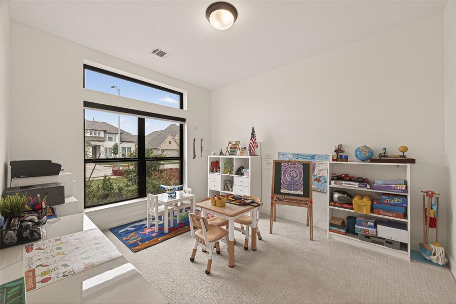 This bright, spacious room is designed as a children's playroom or learning area. It features large windows for natural light, white walls, and carpeted flooring. Equipped with shelves, a small table, and educational toys, it's ideal for a family setting.