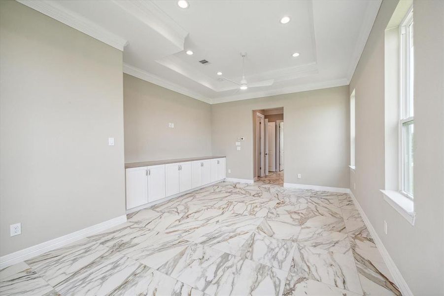 This Master Bedroom features elegant marble flooring with a unique vein pattern, a row of white built-in cabinets for ample storage, tray ceilings with recessed lighting, and large windows for natural light.