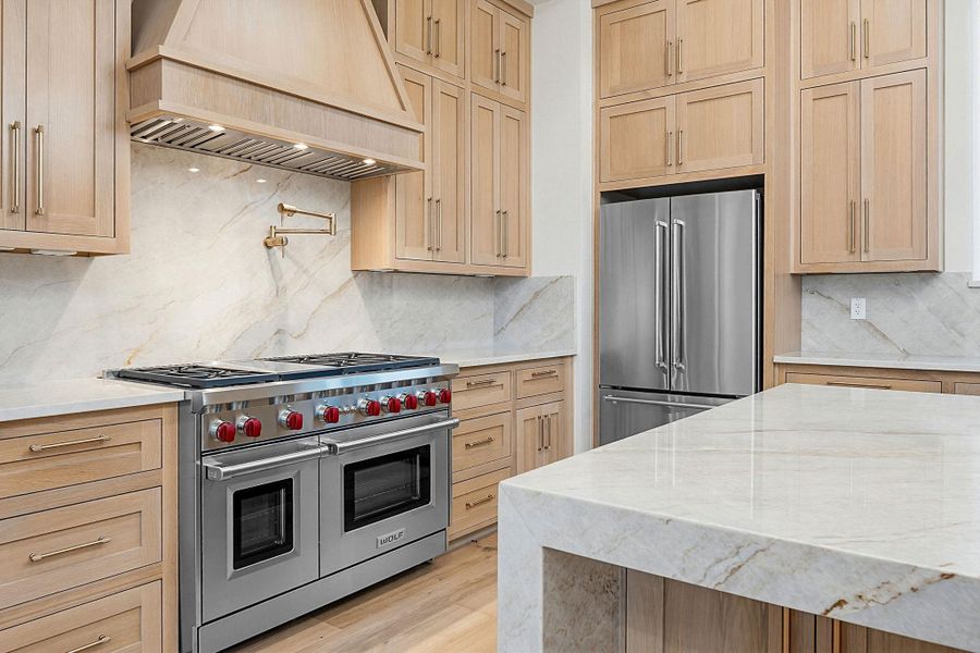 Gourmet kitchen featuring a professional-grade stainless steel range with double ovens, a matching range hood, a stainless steel refrigerator, light wood cabinetry, and stone countertops and backsplash