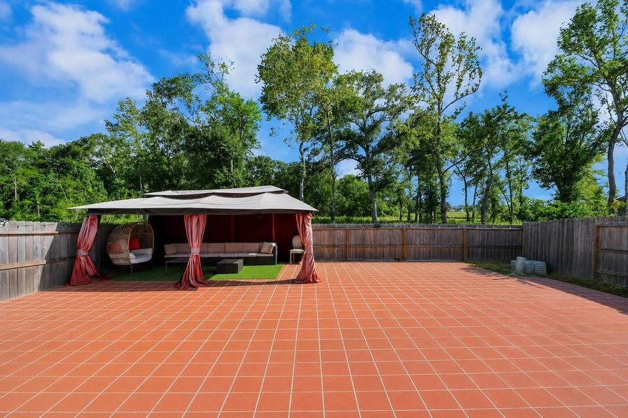This photo features a spacious backyard with a large tiled patio area, surrounded by a wooden fence. There's a cozy gazebo with seating, perfect for outdoor relaxation and entertaining, with lush trees providing a serene backdrop. This photo features a spacious backyard with a large tiled patio area, surrounded by a wooden fence. There's a cozy gazebo with seating, perfect for outdoor relaxation and entertaining, with lush trees providing a serene backdrop.