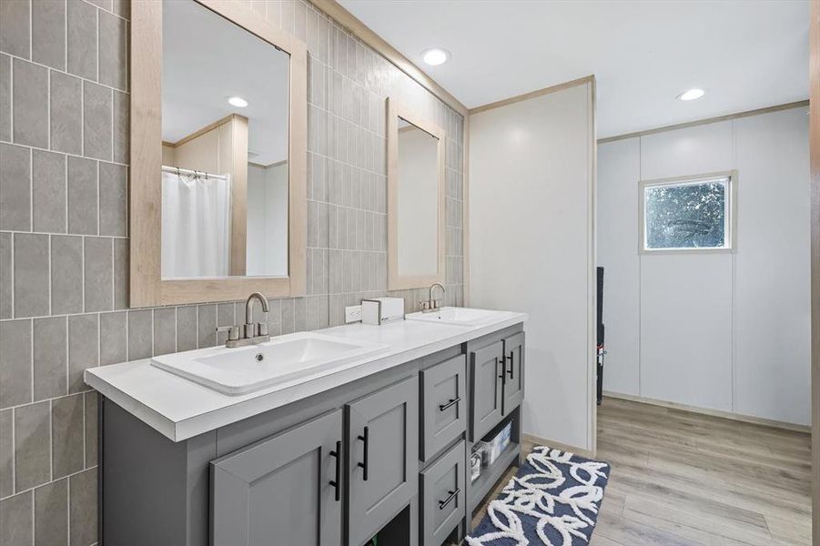 Full bath with double vanity, light wood-type flooring, tile walls, recessed lighting, and a shower with shower curtain Full bath with double vanity, light wood-type flooring, tile walls, recessed lighting, and a shower with shower curtain