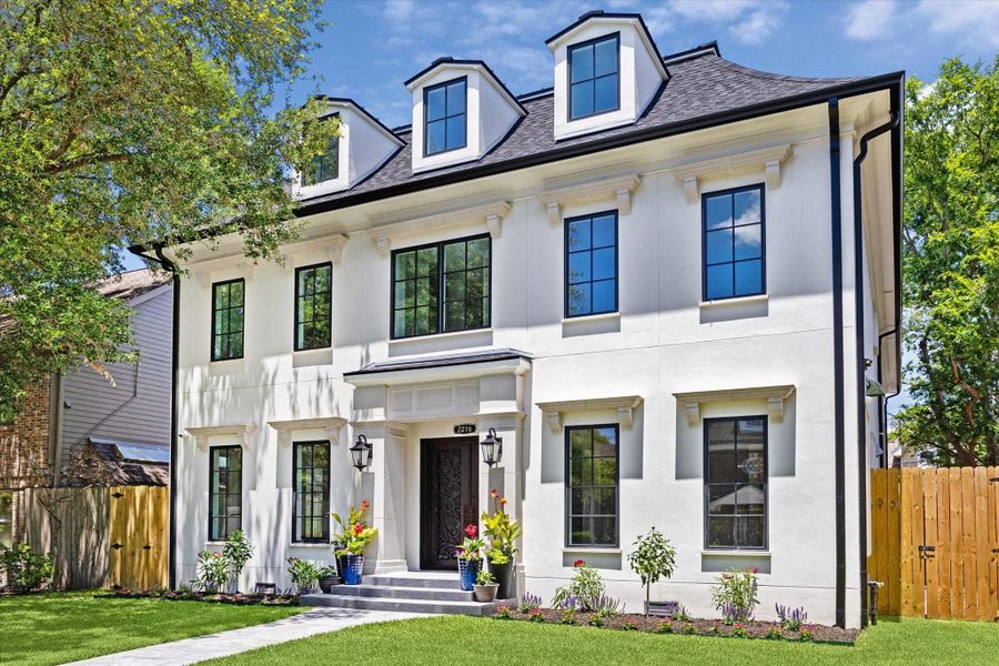 This elegant three story home features a classic white exterior with black-framed windows and dormer accents. It has a manicured front lawn, tasteful landscaping, and a welcoming entrance with potted plants and lantern-style lighting.