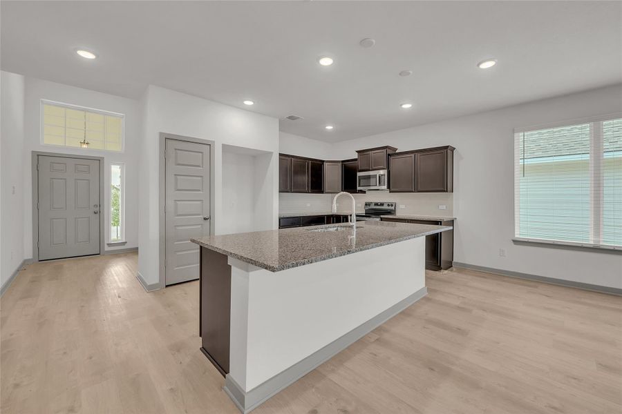 Kitchen with dark brown cabinetry, light stone counters, a center island with sink, healthy amount of natural light, and recessed lighting