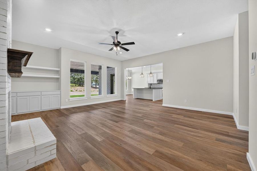 Spacious living area featuring wood-finish flooring, a white brick fireplace with a dark wood mantle, and built-in cabinetry with open shelving Spacious living area featuring wood-finish flooring, a white brick fireplace with a dark wood mantle, and built-in cabinetry with open shelving