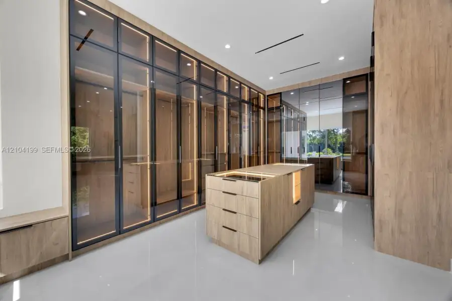 Custom storage system featuring floor-to-ceiling cabinetry with glass-panel doors, integrated LED lighting, central built-in island with storage drawers, wood-finish cabinetry, and high-gloss flooring