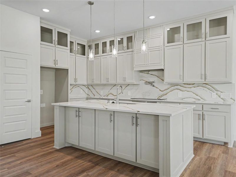 Kitchen featuring light-toned cabinetry, a spacious island with a sink, elegant pendant lighting, a patterned backsplash, and wood-finish flooring