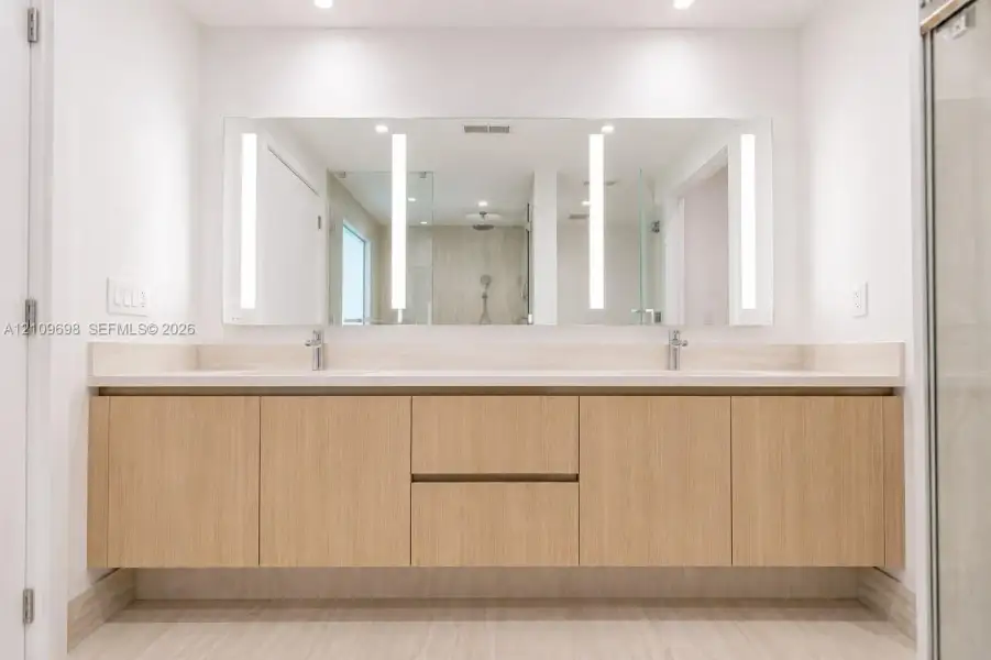 Floating dual-sink vanity featuring light wood-finish cabinetry, solid surface countertop, integrated LED-lit vanity mirror, and polished chrome fixtures