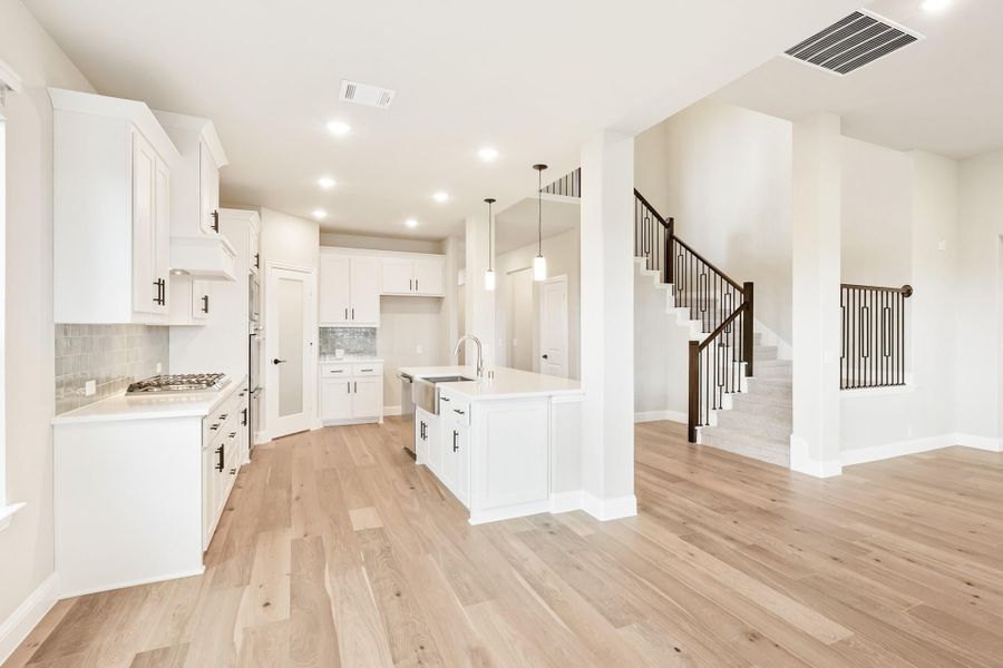 Open kitchen with white cabinets, center island, pendant lights, and light hardwood floors, with staircase visible nearby.