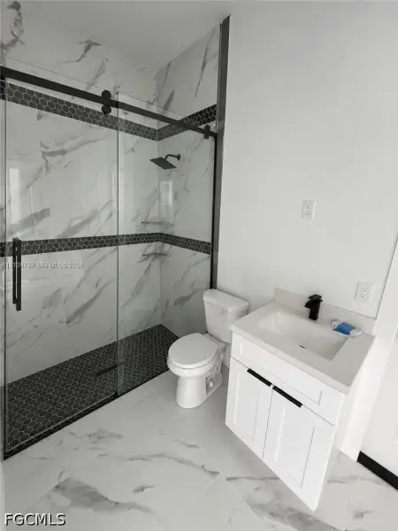 Walk-in shower with floor-to-ceiling stone-pattern tile, hexagonal mosaic tile flooring, glass enclosure with matte black hardware, white vanity cabinet with light-toned countertop, and matte black plumbing fixtures