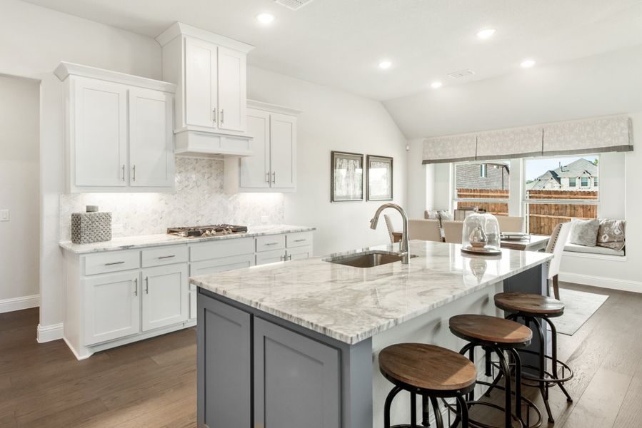 Kitchen with white cabinets, marble backsplash, gray island with sink, and three bar stools on dark hardwood floors