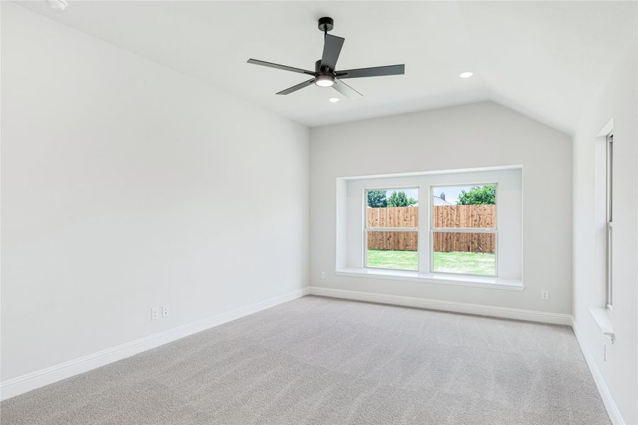 Empty bedroom with vaulted ceiling, black ceiling fan, carpet flooring, and a large window overlooking a fenced backyard